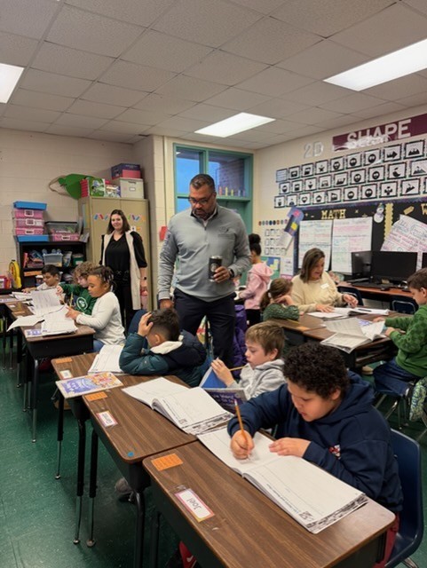 A classroom with children seated at desks, writing in their notebooks. A man in a gray shirt stands at the front.