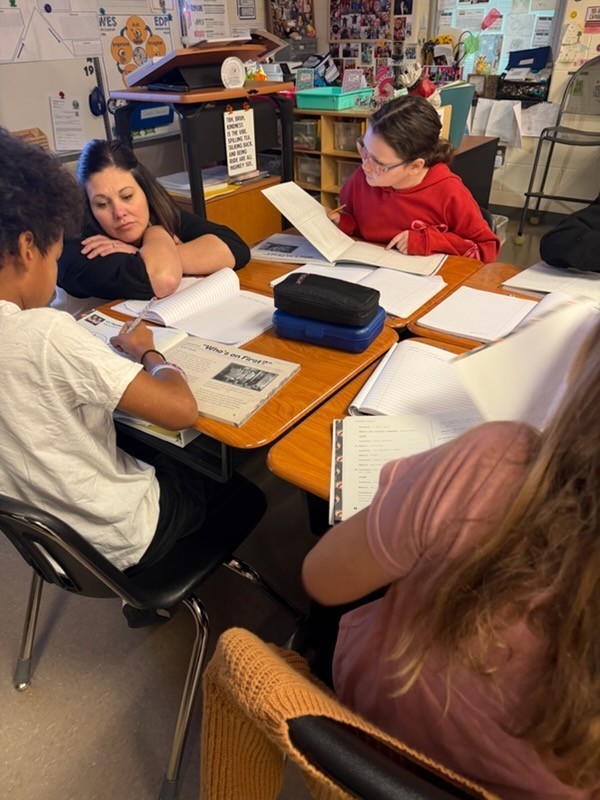 Four people sit at a desk. One person wears glasses. Two people have papers. A woman looks down.