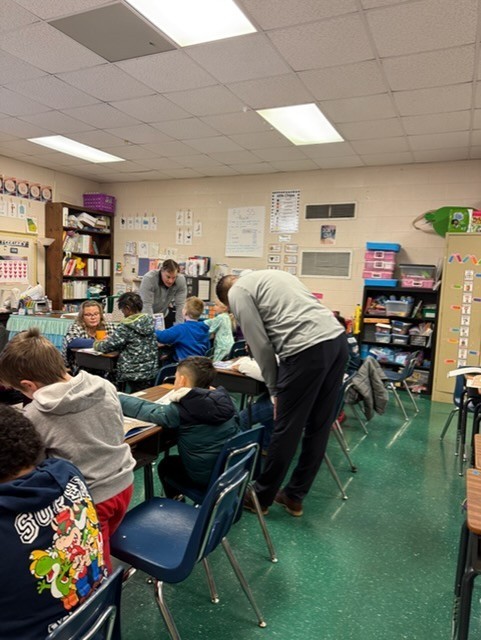 A classroom with green floor and ceiling lights. A man bends over a student's desk. Students sit at their desks.