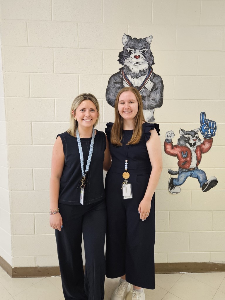 Two women in navy jumpsuits stand next to a white wall with drawings of a cat and a raccoon.
