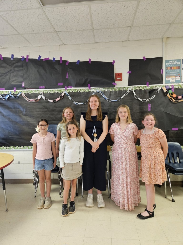 Six children and an adult woman stand in a classroom. They are wearing casual clothing and shoes.