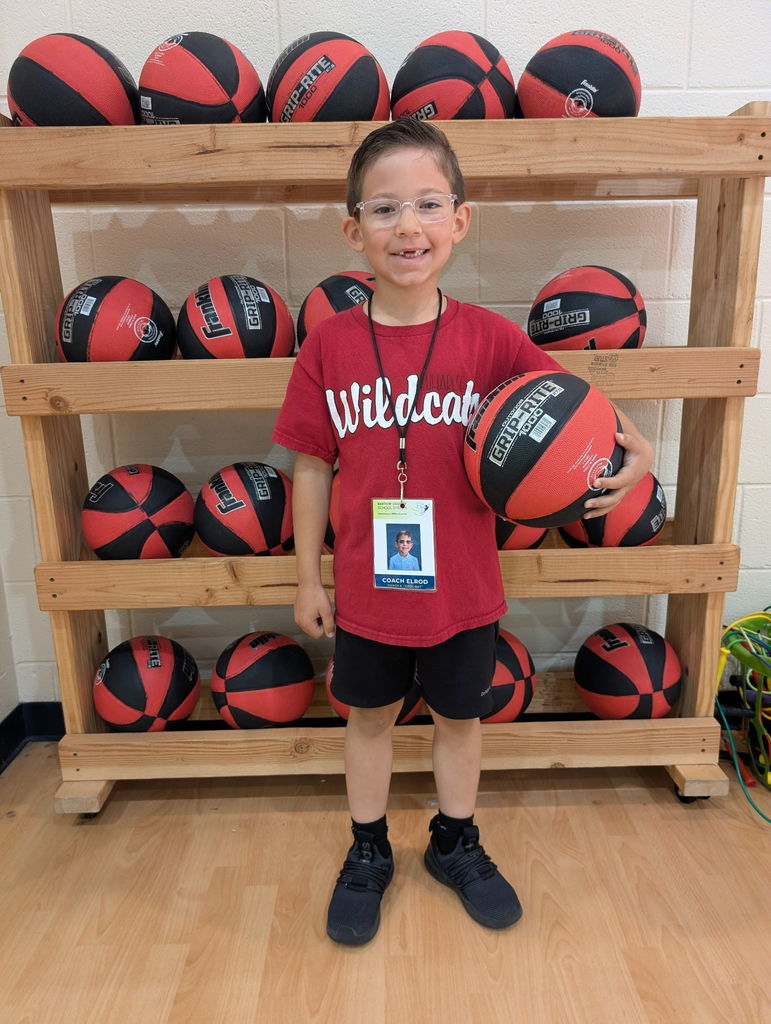 A child stands in front of a wooden shelf with multiple basketballs. They wear a red shirt and glasses.  He is wearing a burgundy shirt that says "Wildcats".