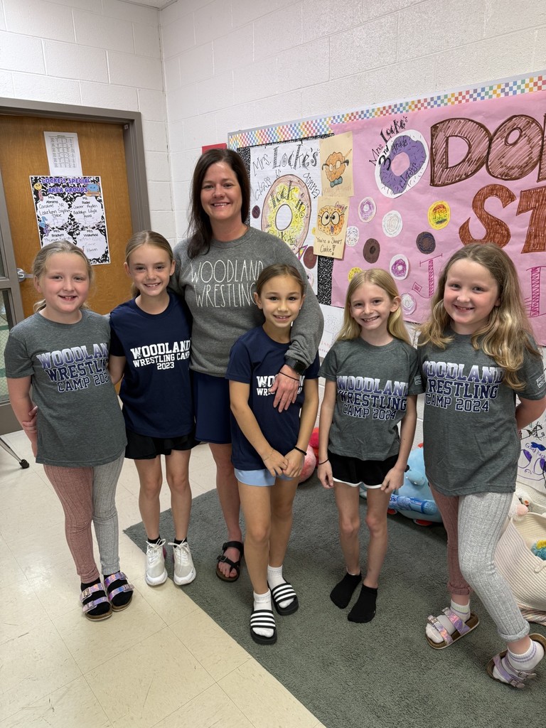 Six young girls in matching grey t-shirts, posing for a photo with a woman, standing in a room with a bulletin board.  They are all wearing shirts that say "Woodland Wrestling".