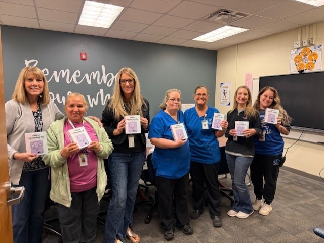 A group of women in blue scrubs and one in a pink shirt stand in a room, holding certificates. Behind them is a gray wall with a blackboard.