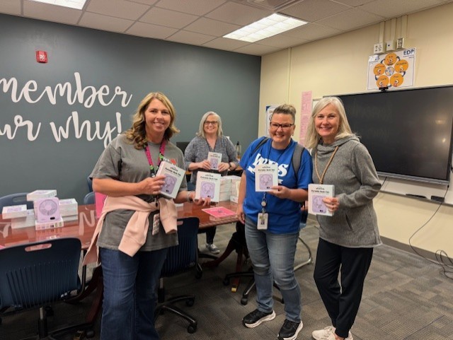Four adults stand in a room with a table, holding small items. One wears a blue shirt and sneakers. A wall reads "member our why!"