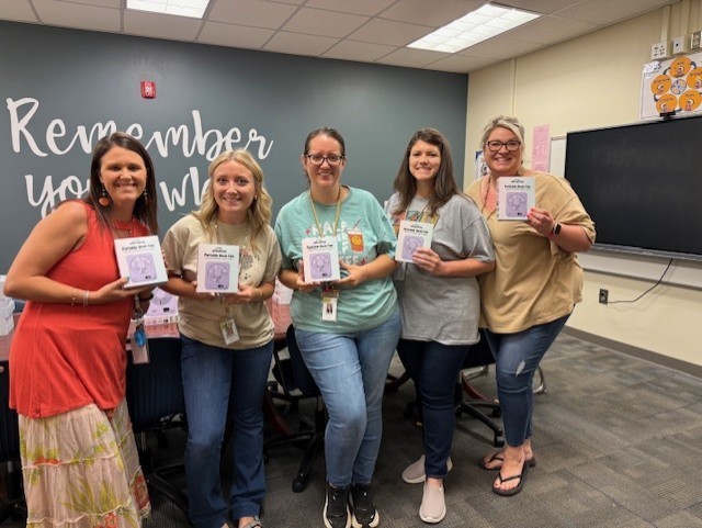 Five women in a classroom holding certificates, standing next to each other, with a wall that says "Remember you".