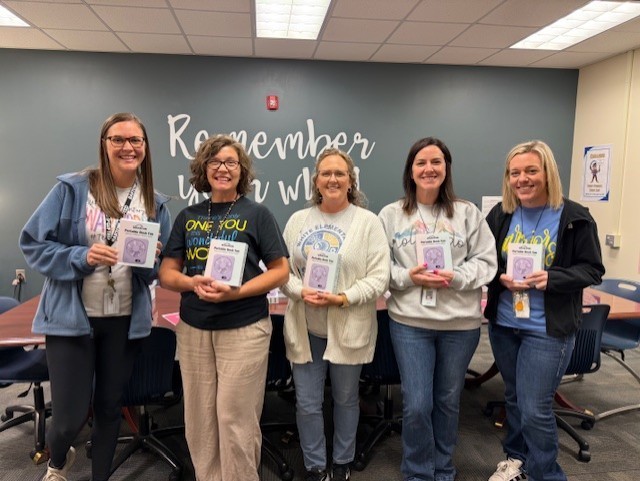 Five women stand together holding books. A wall behind them reads "Remember". A table with chairs is in front.