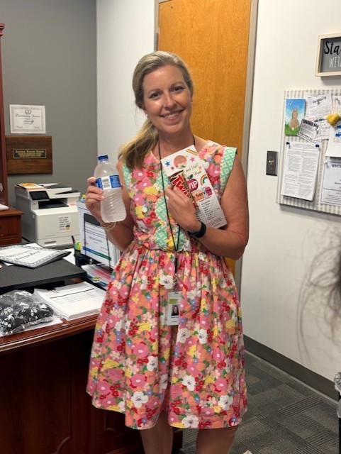 A woman in a floral dress stands in an office, holding a box and a water bottle. Behind her, a desk has papers and a printer. Bulletin board with papers.