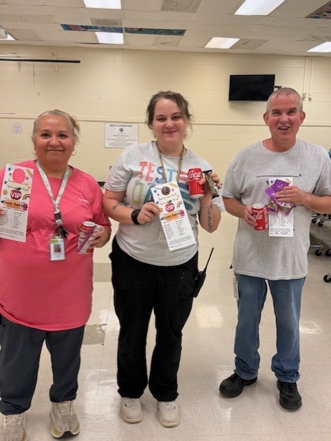 Three people in a room with white walls, smiling and holding cups. One wears a pink shirt, another a gray shirt, and the third a black shirt.
