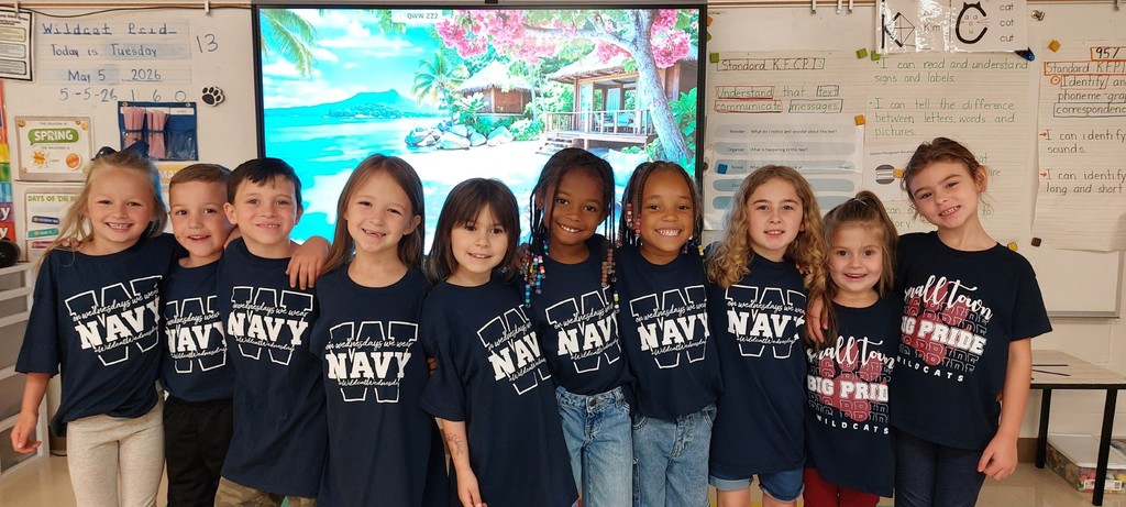A group of children in navy blue t-shirts stand close together in a classroom. Behind them, a screen displays an image.  On their shirts is the word "Navy" and "Big Pride:.