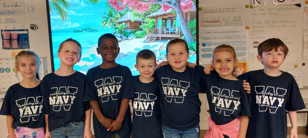 Six children in navy blue T-shirts, one with pink shorts, standing in front of a large screen.  On their shirts is the word "Navy".