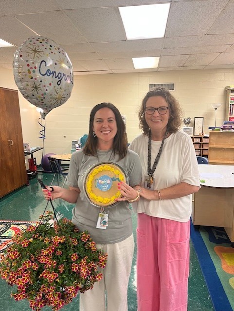 Two women stand in a classroom. One holds a balloon and a plant, and the other holds a plate.