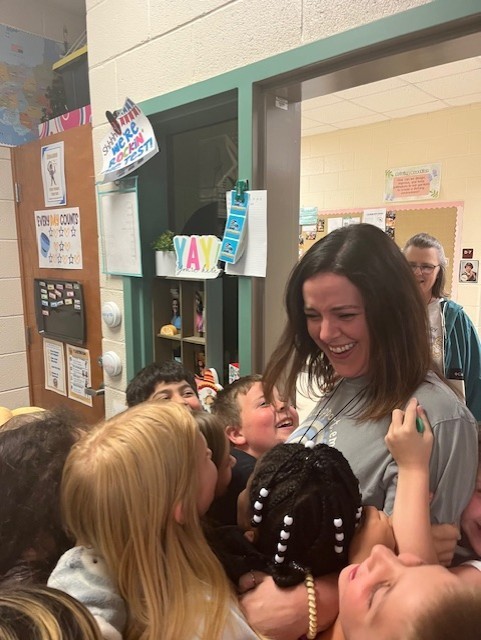 A woman smiles as children gather around her in a classroom, with bulletin boards and a shelf visible in the background.