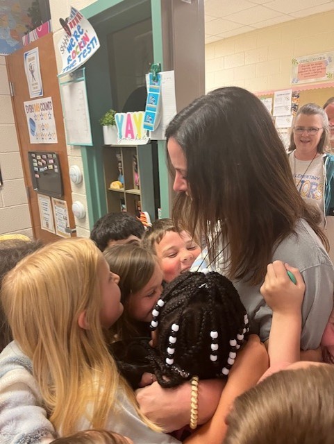 A woman with long dark hair is surrounded by children. Some children are hugging her while others watch.