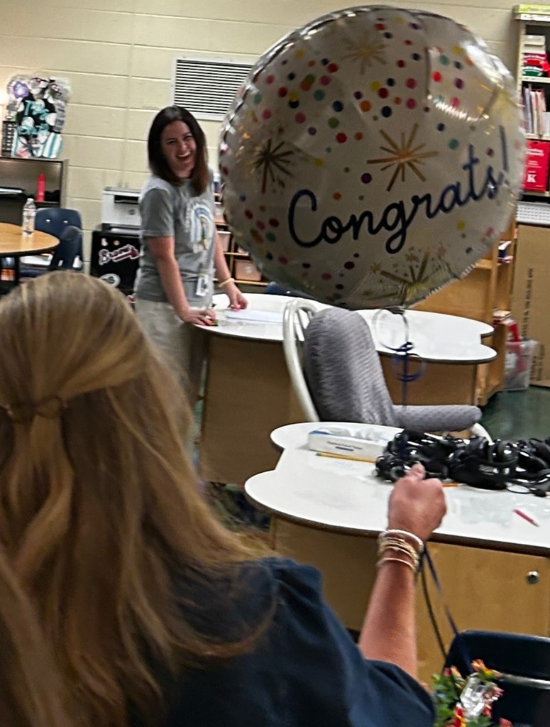 Two women in a classroom. One holds a balloon with "Congratulations" written on it. Background includes tables and chairs.