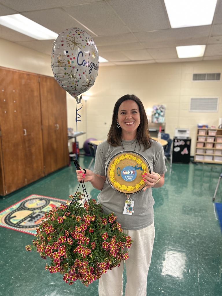 A woman stands in a room, holding a balloon that says "Congrats" and a round award, with a bouquet of flowers.