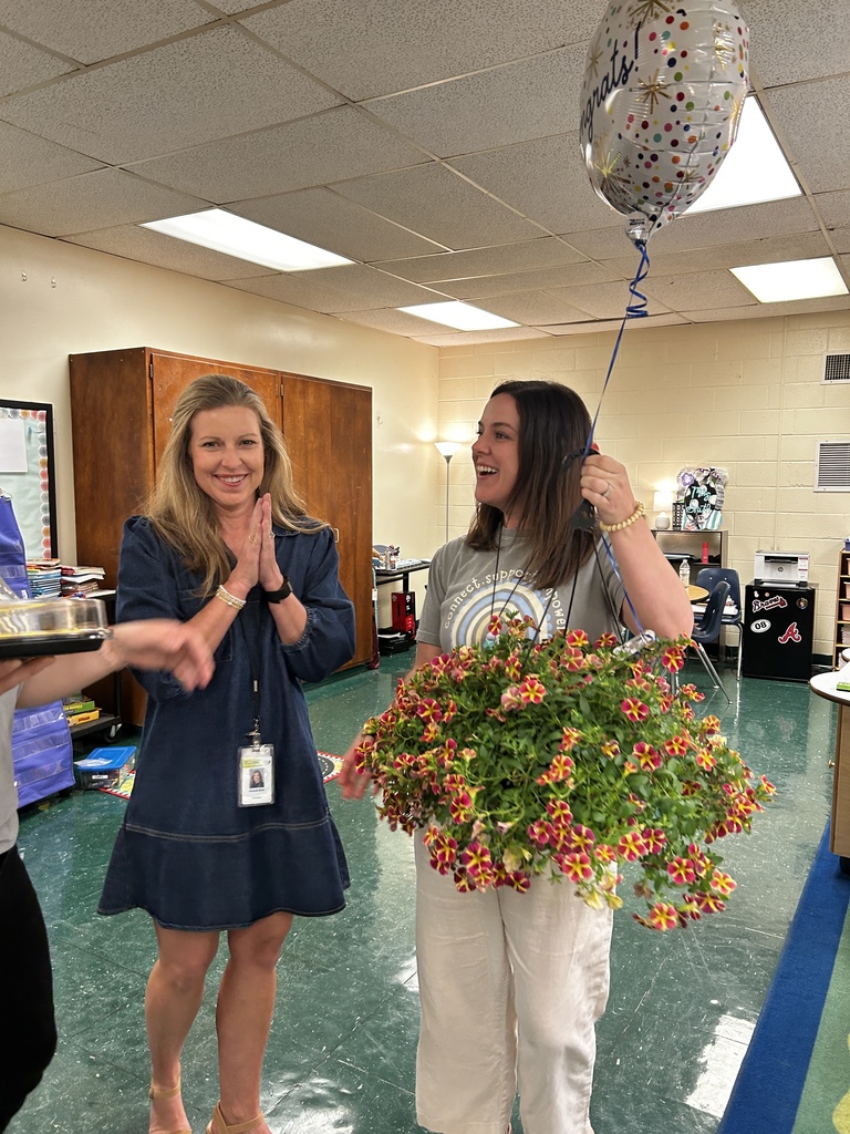 Two women standing in a room with balloons and flowers. One holds flowers, and the other holds a balloon.