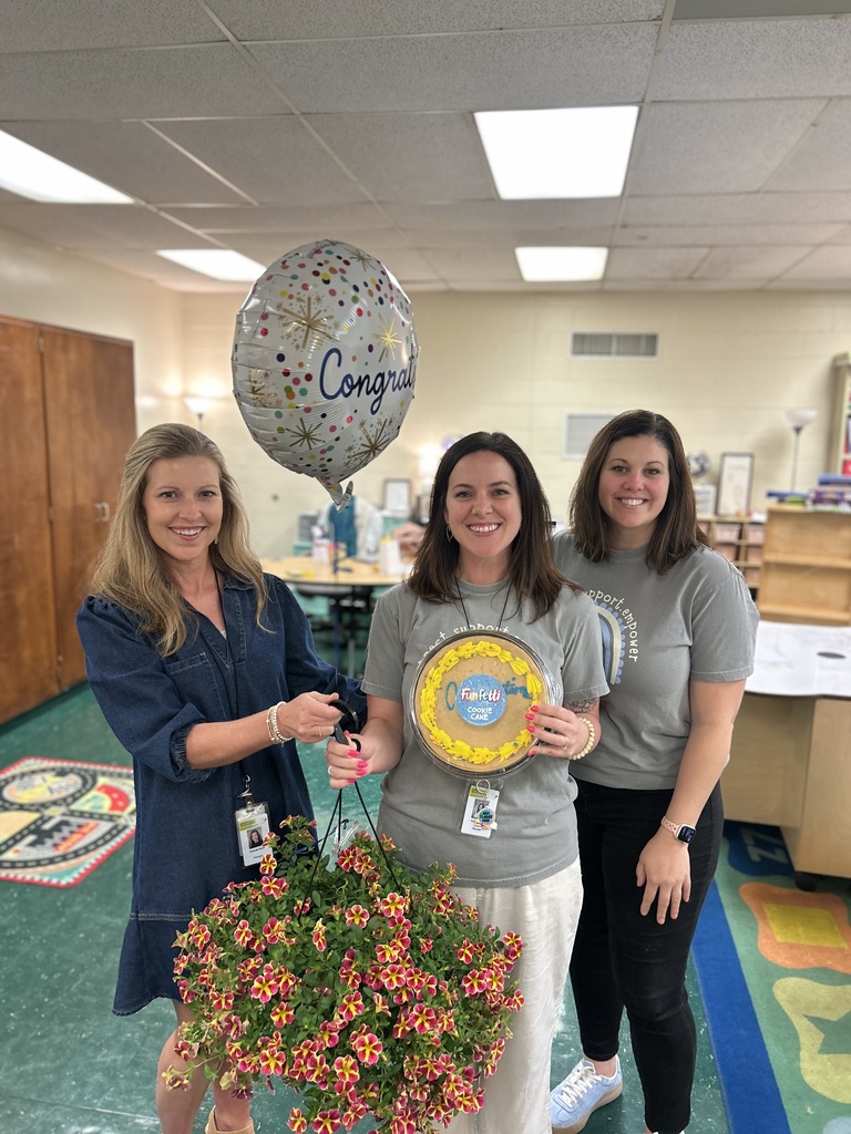 Three women stand next to each other in a room, one holding a balloon and another a bouquet.