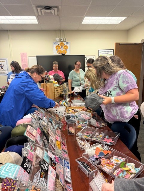Several people are in a room examining items at a table with various items. One person is wearing a blue jacket.