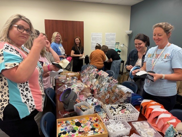 Several women, wearing eyeglasses and watches, are standing around a table filled with various items.