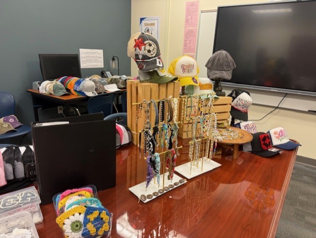 Display of various hats and necklaces on a desk in a room with a large screen TV.