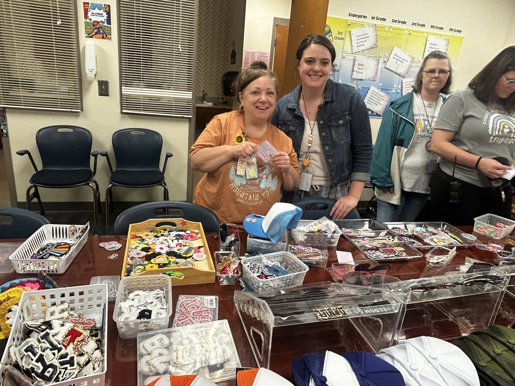 Three women at a table with various craft supplies, including containers, baskets, and hats.