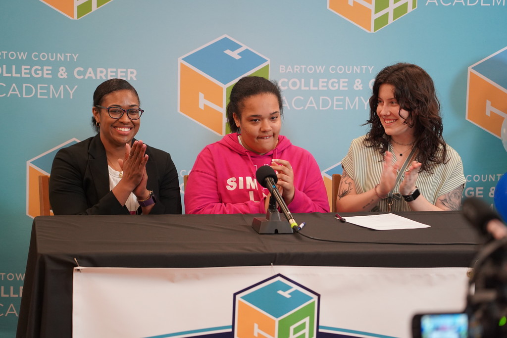 Three women, one in a pink hoodie, sit behind a table with microphones. They appear to be at a press conference.