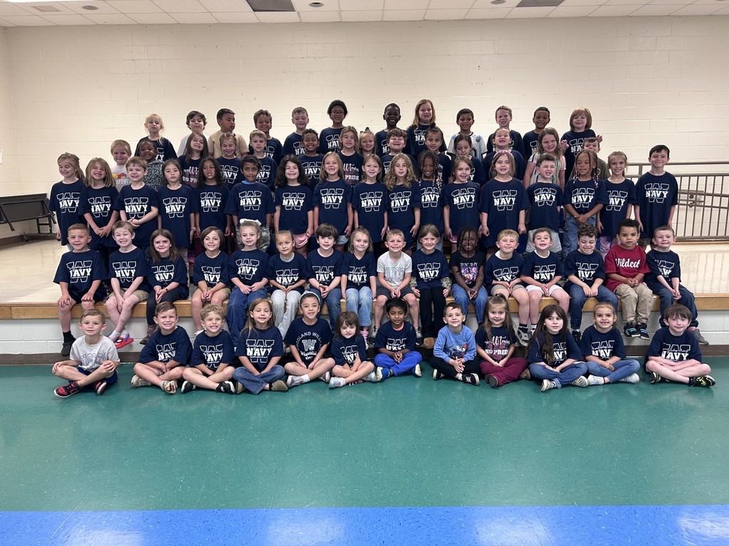 A group of children in navy blue shirts with W's on them, posing for a photo in a lunchroom.