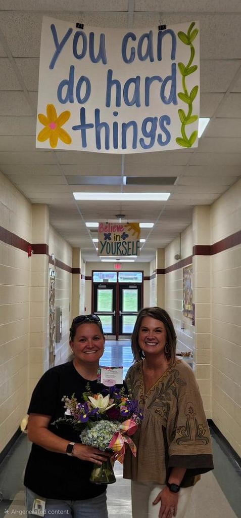 Two women stand in a school corridor with a motivational banner above. One holds flowers, both smiling.