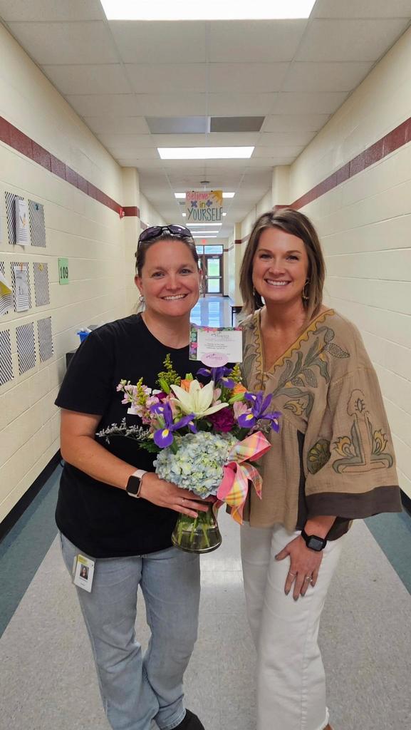 Two women stand in a hallway, one holding flowers, both smiling, with a school hallway background.