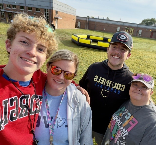 Four people standing in a field in front of a building. They are smiling. One wears a red shirt, another a white sweatshirt, and two others wear black and gray tops.
