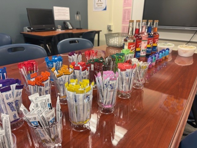 A wooden table has many plastic cups filled with sugar packets, candies, and other colorful items. Behind the table is a monitor and a desk.