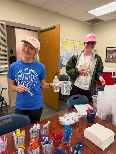 Two women, one holding a bottle, smile in a room with a table full of cups and snacks.
