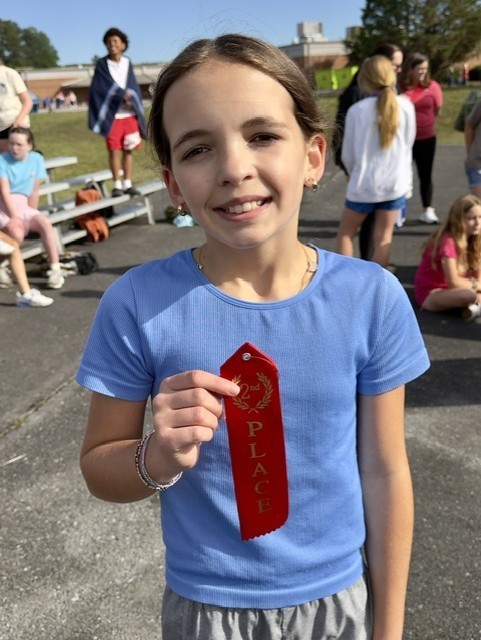 A young girl in a blue shirt holds a red ribbon labeled "PLACE." She smiles in an outdoor area.