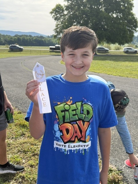 Boy holding a white award ribbon with a blue shirt reading "Field Day" outside.