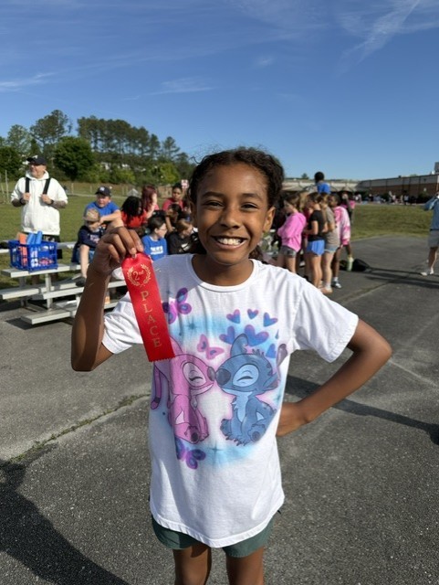 Child holding a red ribbon and smiling at an outdoor event. Behind, people are standing and sitting on benches.