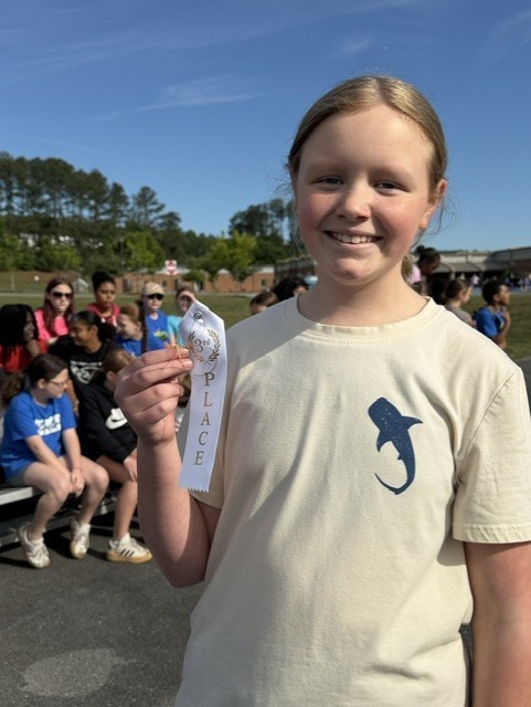 A young girl displays a medal while standing in a school parking lot, with a crowd in the background.