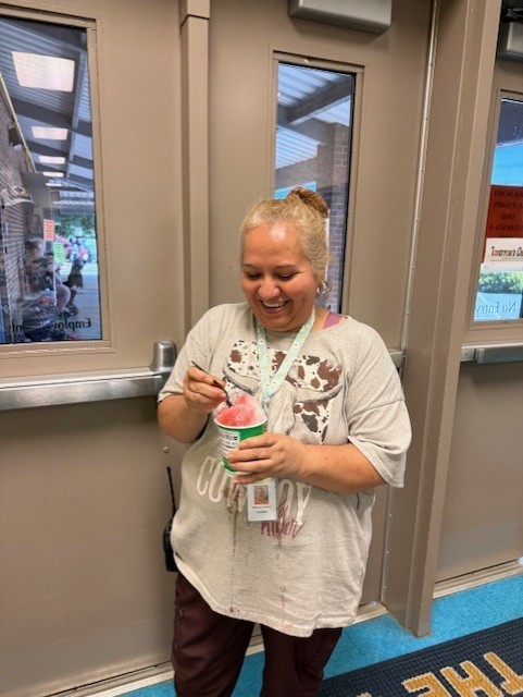 A woman with a bun hairstyle is holding a cup of pink ice cream, smiling in a building.