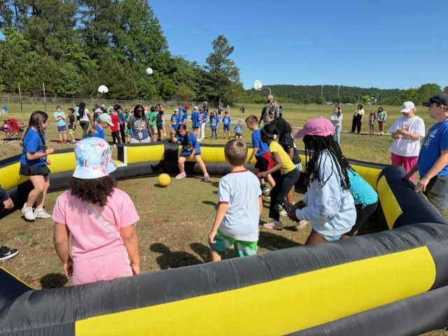 Kids playing in a large inflatable obstacle course on a grassy field, with onlookers standing nearby.