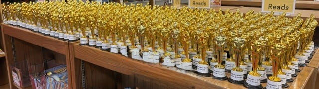 Many golden trophies arranged in rows on wooden shelves. Each trophy has a white base.