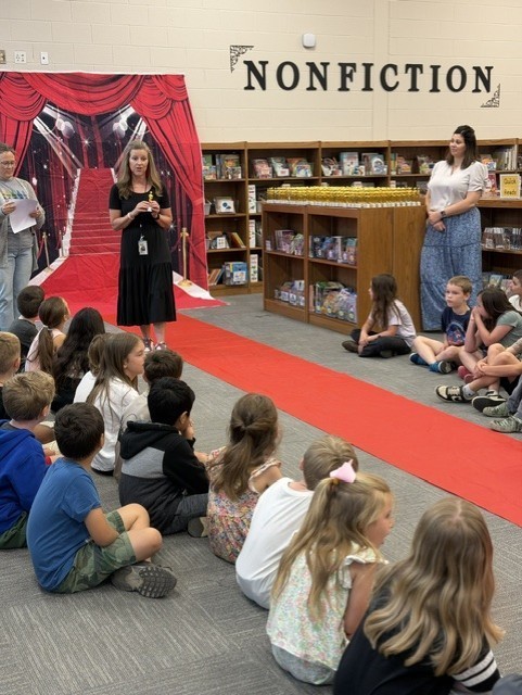 Children seated on a floor, facing adults on a red carpet with a "Nonfiction" sign in a library.