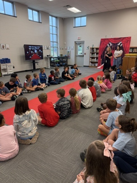 Children sit on the floor in a classroom, facing a stage with a red curtain. Some are on a red carpet.