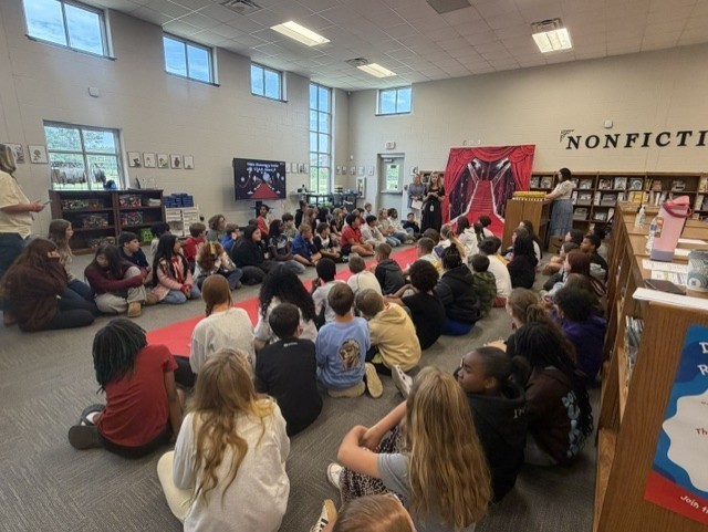 Large group of children seated in a circle on the floor in a school library.