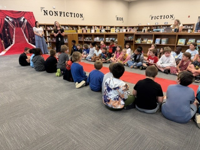 Kids sit on a carpeted floor in a library, listening to a woman speak. Shelves of books line the background.