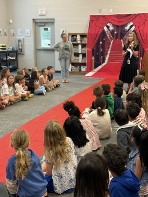 Group of students sit on floor in front of two women on a stage. Behind them is a large red curtain.
