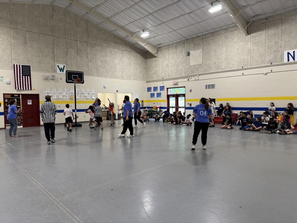 Indoor basketball court with people playing. Some people are sitting on the floor watching. A flag is displayed on the wall.