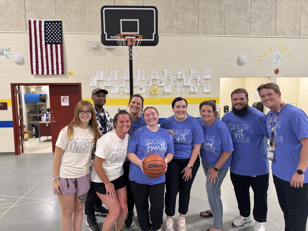 A group of individuals in blue shirts pose for a photo in a gymnasium. A basketball hoop is in the background.