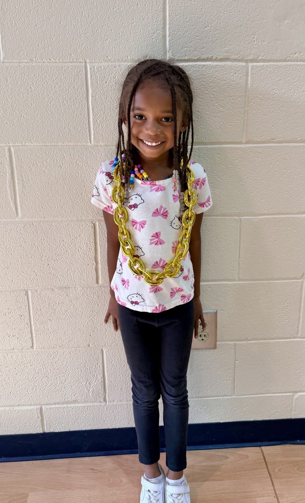 A young girl with braided hair, smiling, wears a yellow necklace. She stands in front of a white brick wall.