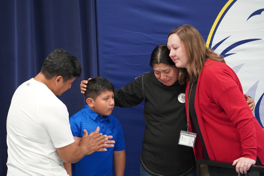 People standing, with a man and woman embracing a child. A man and woman stand behind a blue backdrop with a yellow and white logo.