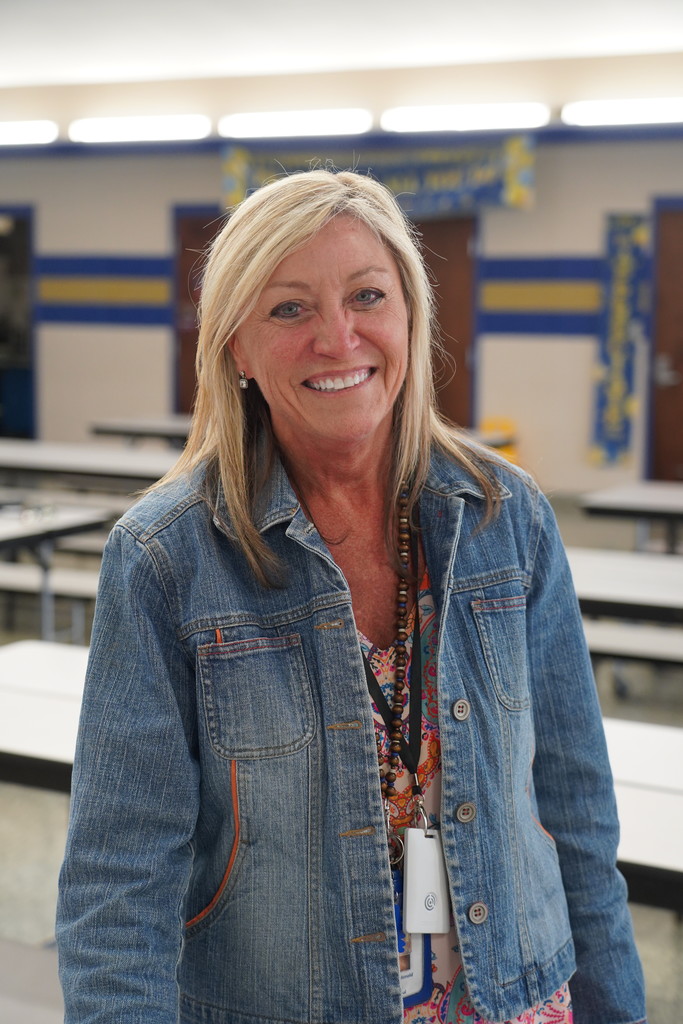 Woman with blonde hair smiling, wearing a denim jacket, and standing in a classroom with tables and chairs.